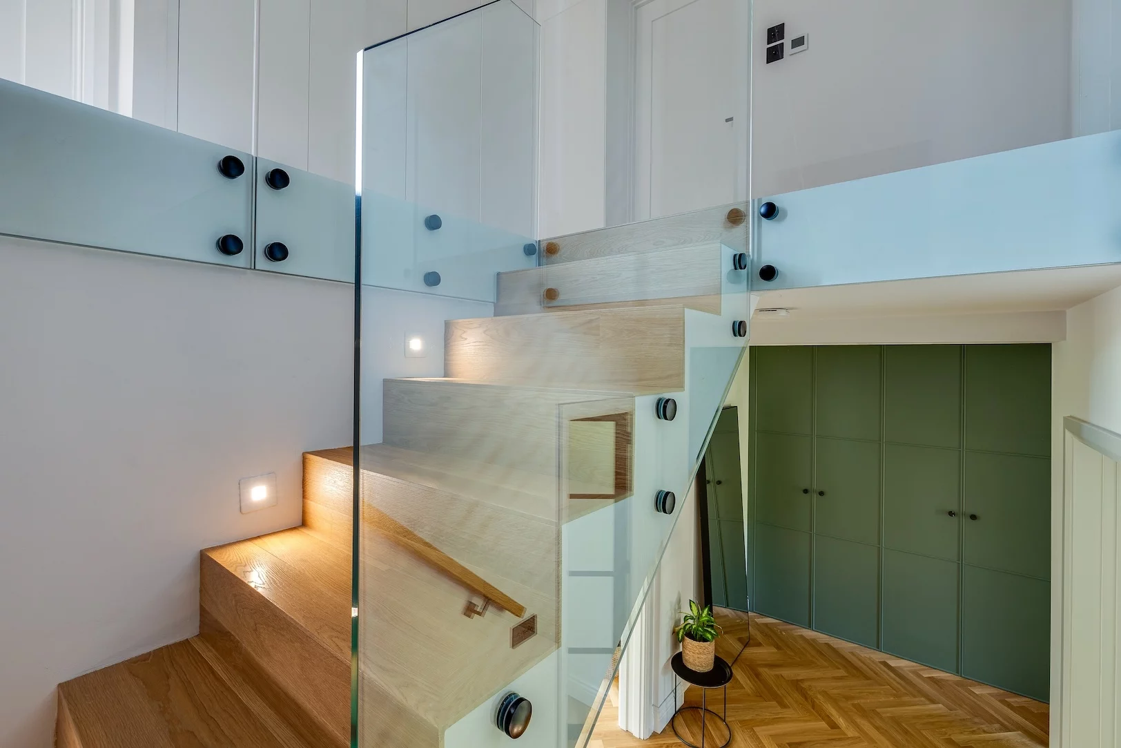 Modern wooden staircase with glass balustrade and black fittings, recessed wall lights and green built-in cupboards below.