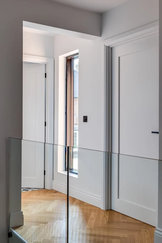 Minimal white hallway with a tall narrow window, glass balustrade and light oak herringbone floorboards.