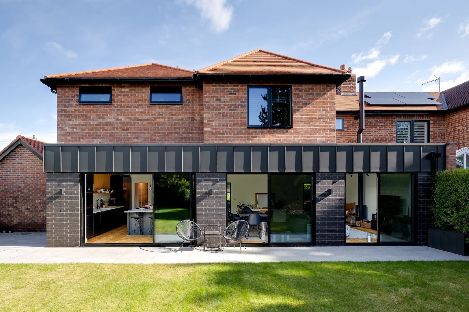 Two-storey brick house with large ground-floor sliding glass doors opening onto a lawn and two black patio chairs.