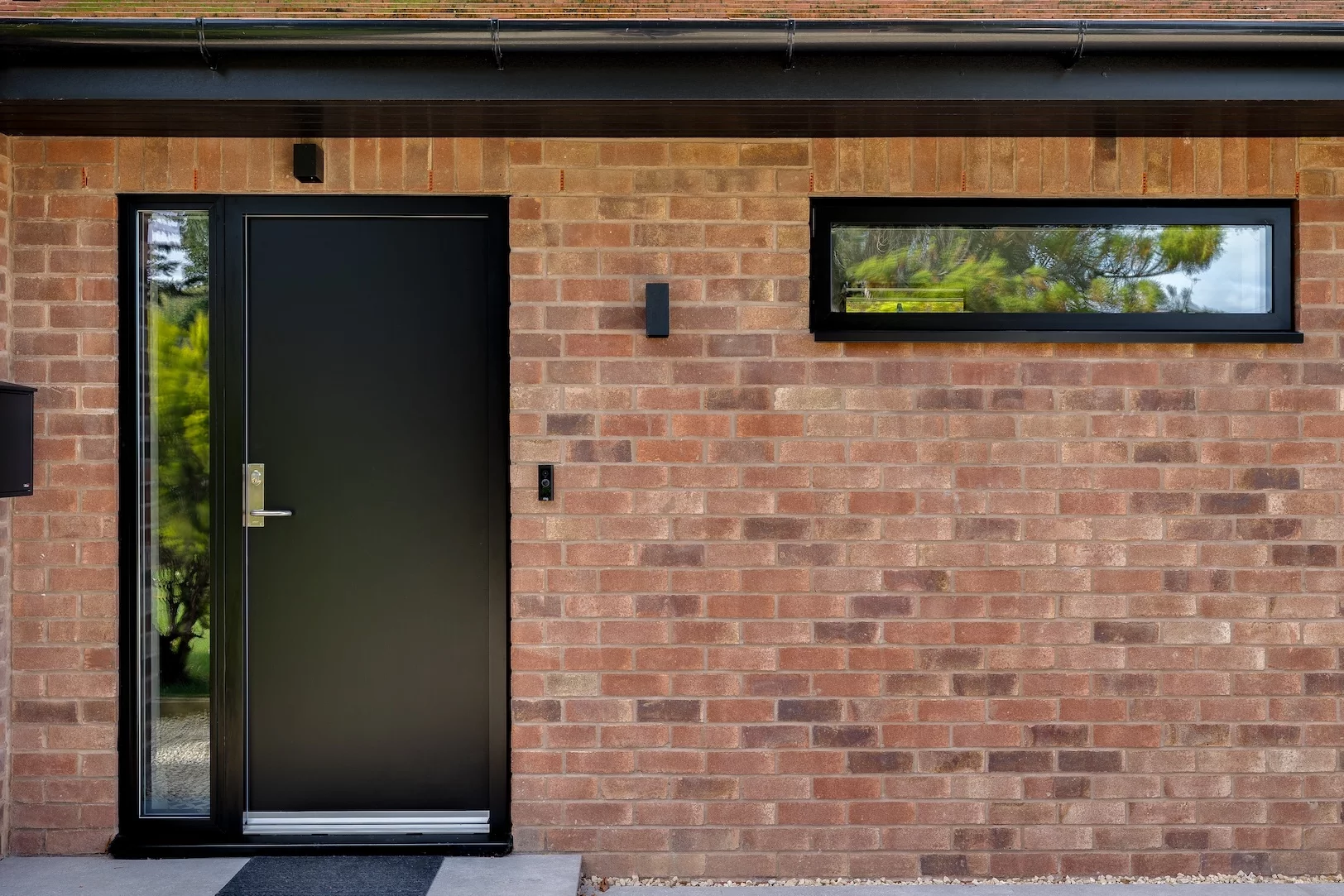 Matte black front door set in red brick wall with narrow sidelights, horizontal window, and modern doorbell light fixture