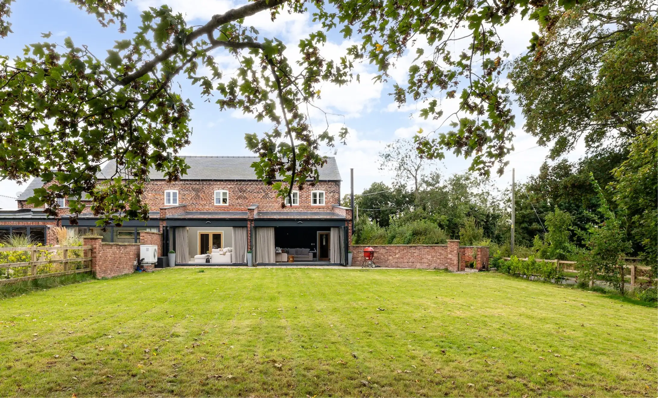 Brick country house with open patio doors, sofas visible inside and a large grassy lawn framed by overhanging tree branches