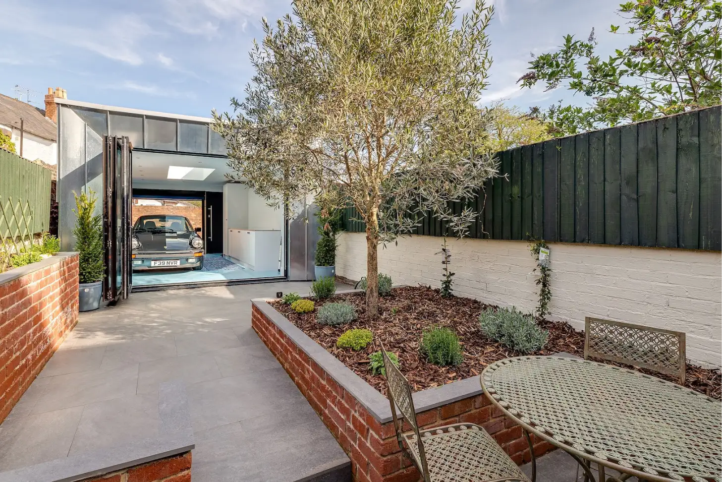 Rear garden with paved patio, raised brick planter and olive tree, metal bistro set, and a modern garage with a parked car.