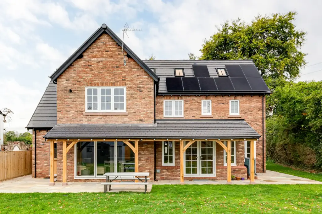 Two-storey redbrick house with slate roof, solar panels on the upper right roof, white-framed windows and a wooden porch over sliding patio doors.