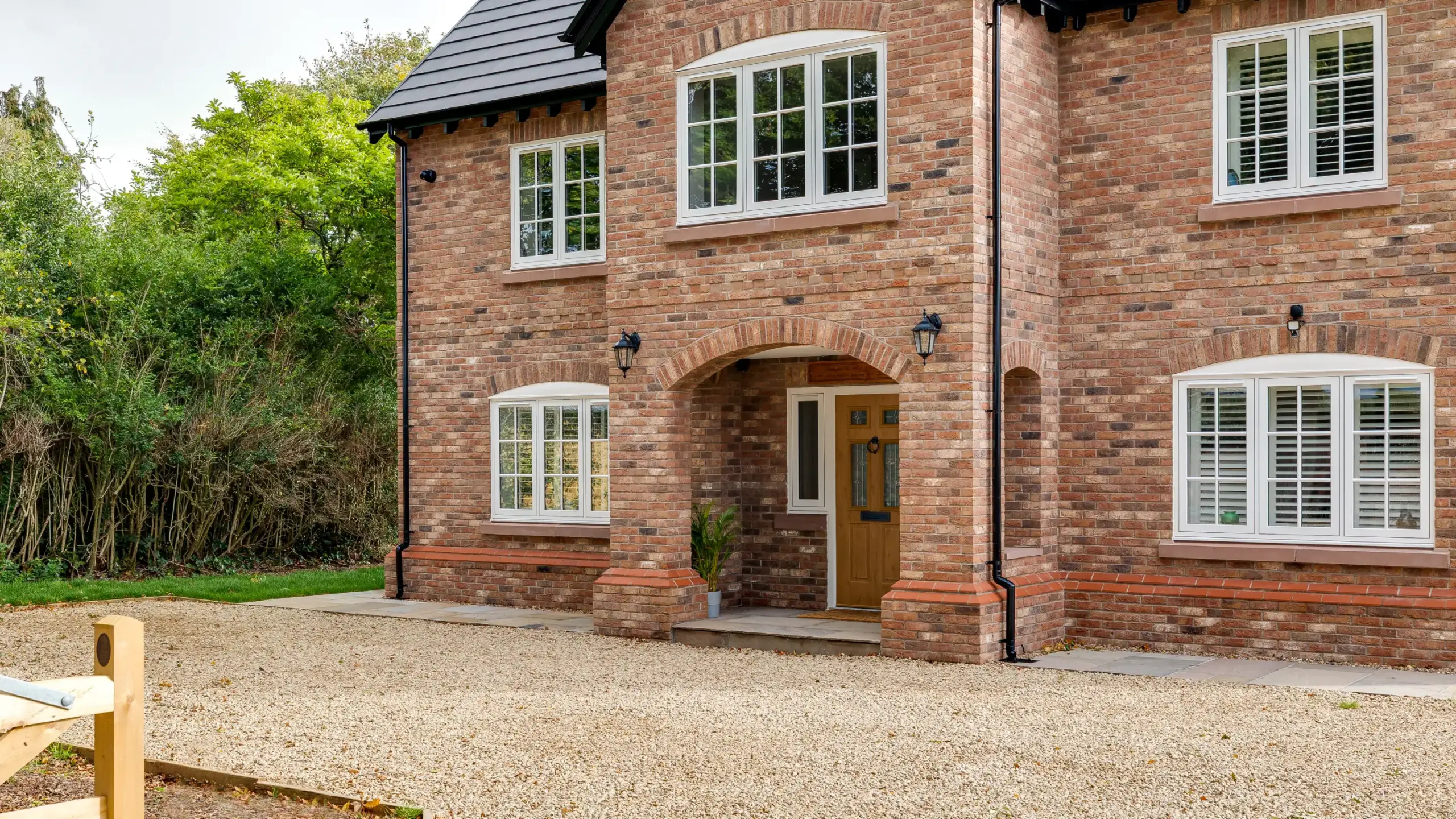 Two-storey redbrick house with white-framed windows, arched porch, wooden front door and gravel driveway surrounded by shrubs
