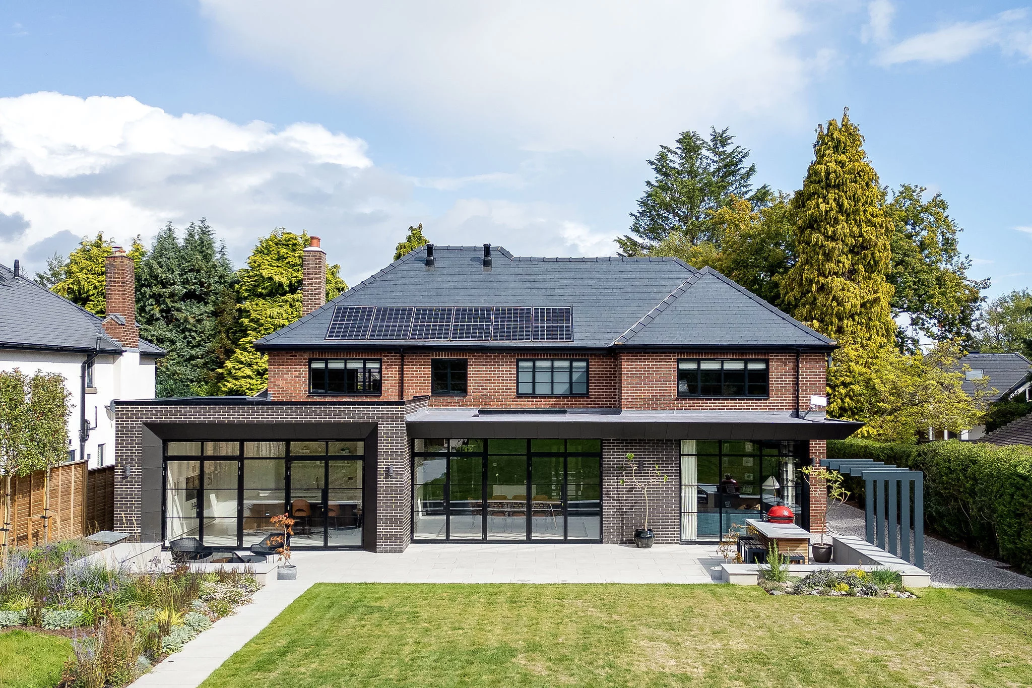 Two-storey brick house with large glass doors, slate roof with solar panels, patio and lawn in front, tall trees behind.