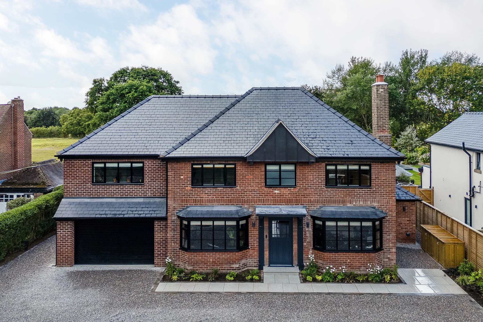 Two-storey red-brick house with dark slate hipped roof, three black-framed windows above bay windows and central dark blue door.