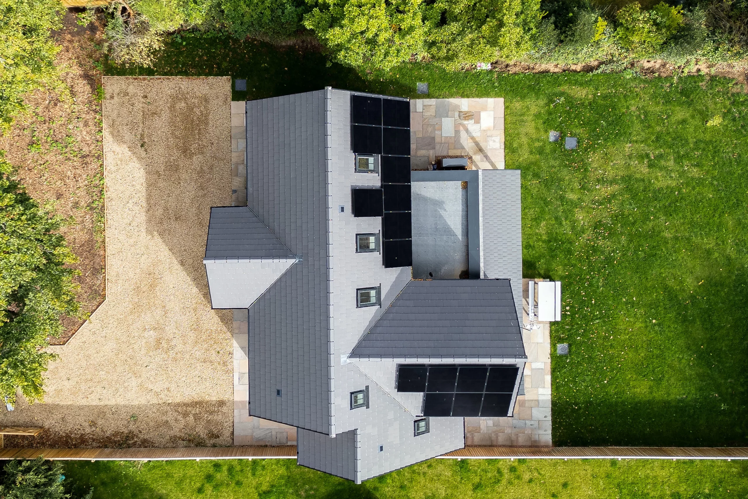 Aerial view of a grey-roofed house with black solar panels, surrounding green lawn on the right and gravel yard on the left.
