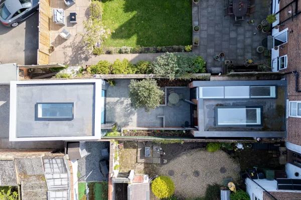 Aerial view of narrow back gardens and patios between terraced houses, showing paved courtyards, planters, lawns and skylights on flat roofs.