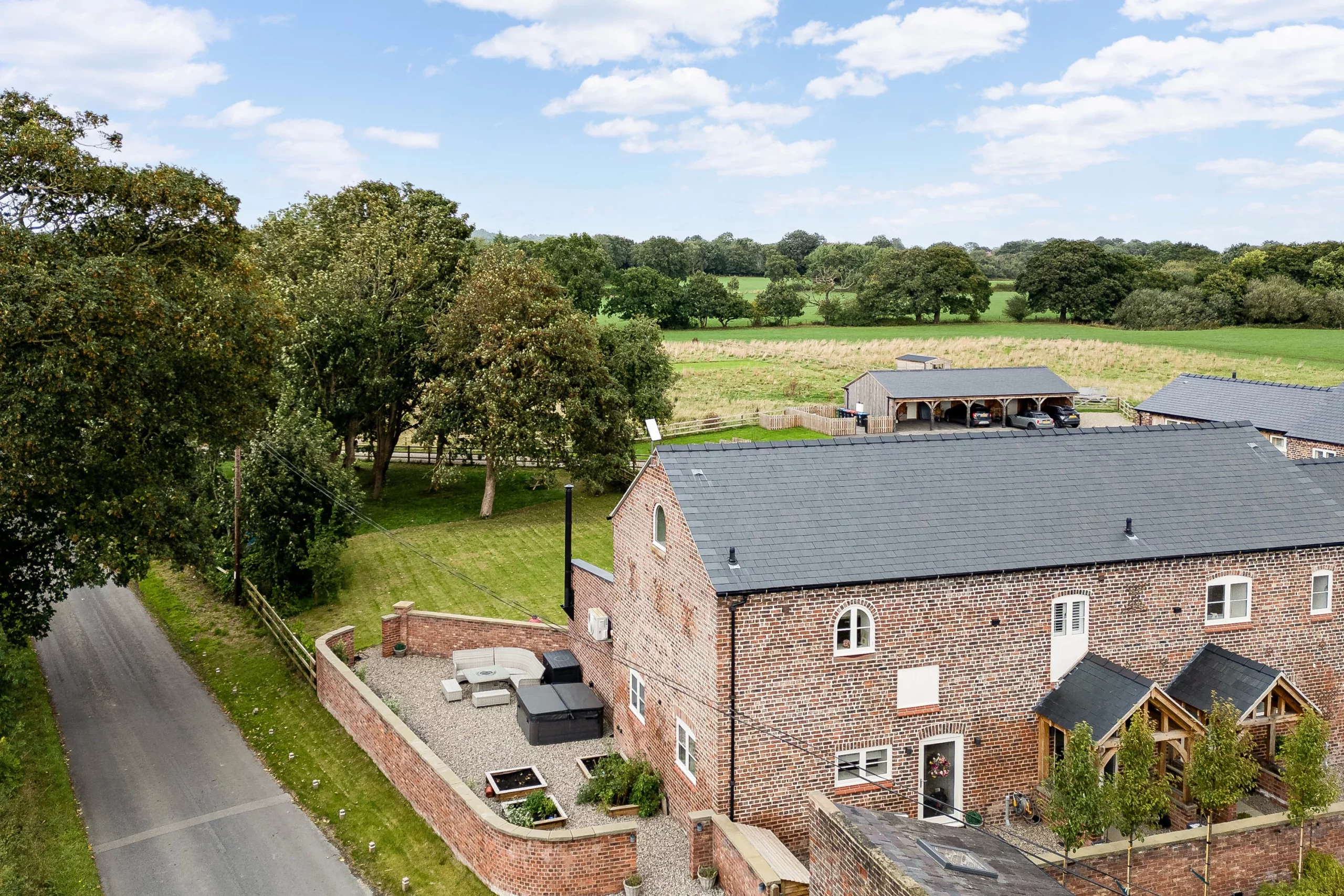 Red-brick farmhouse with slate roof, courtyard seating and carport, surrounded by trees, hedgerow and open green fields under a blue sky
