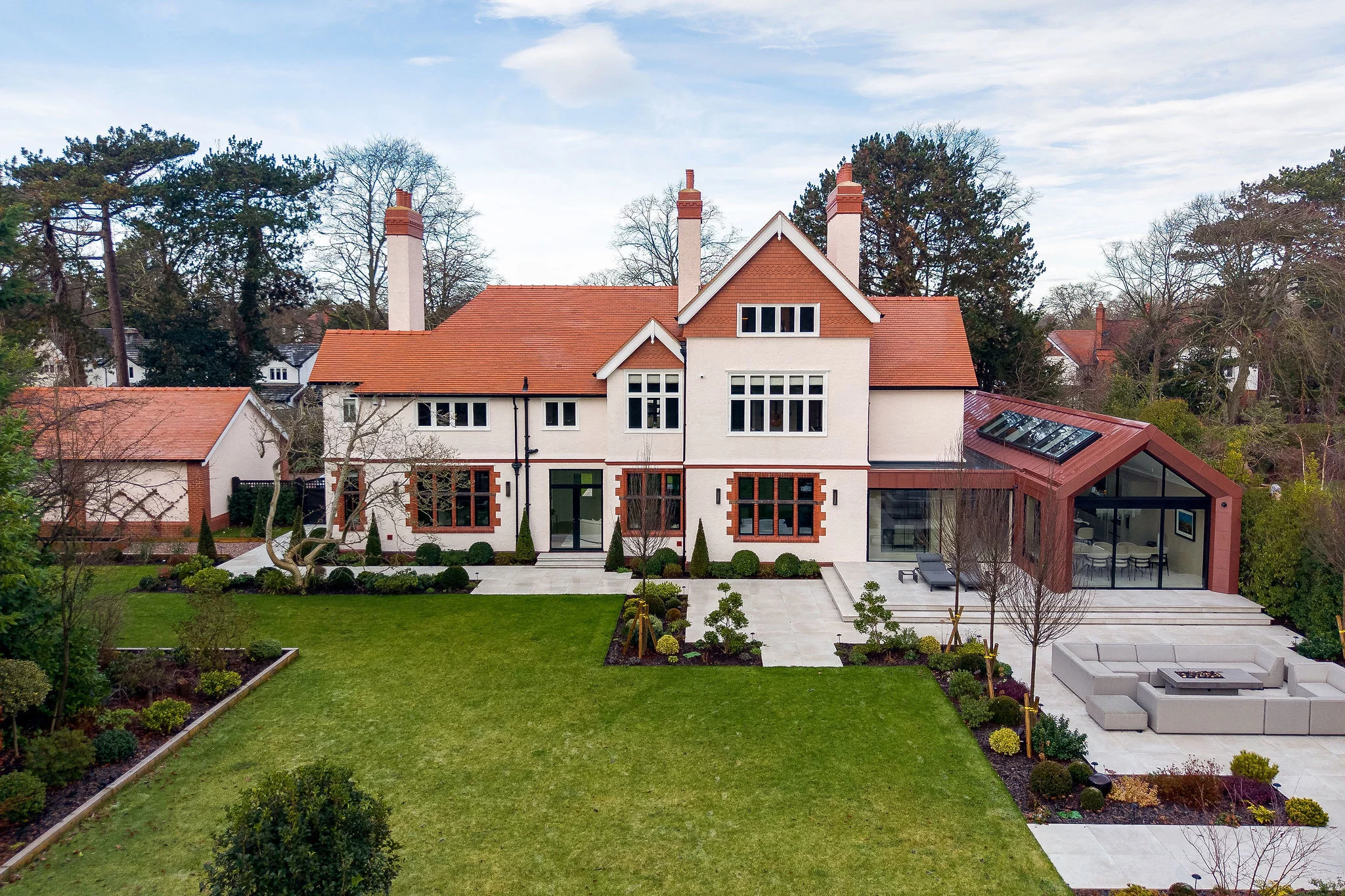 Large two-storey cream house with red tiled roof, chimneys and modern glass extension, surrounded by manicured lawn and patio seating.