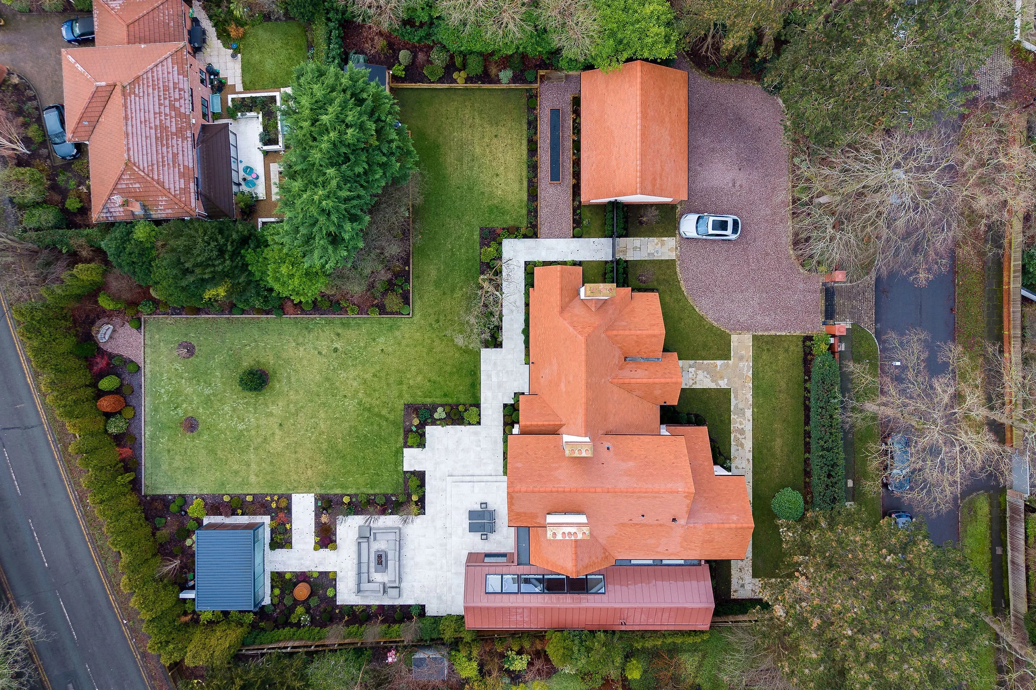 Aerial view of a large suburban house with red roofs, paved patios, driveway with a parked car, and fenced green lawn.
