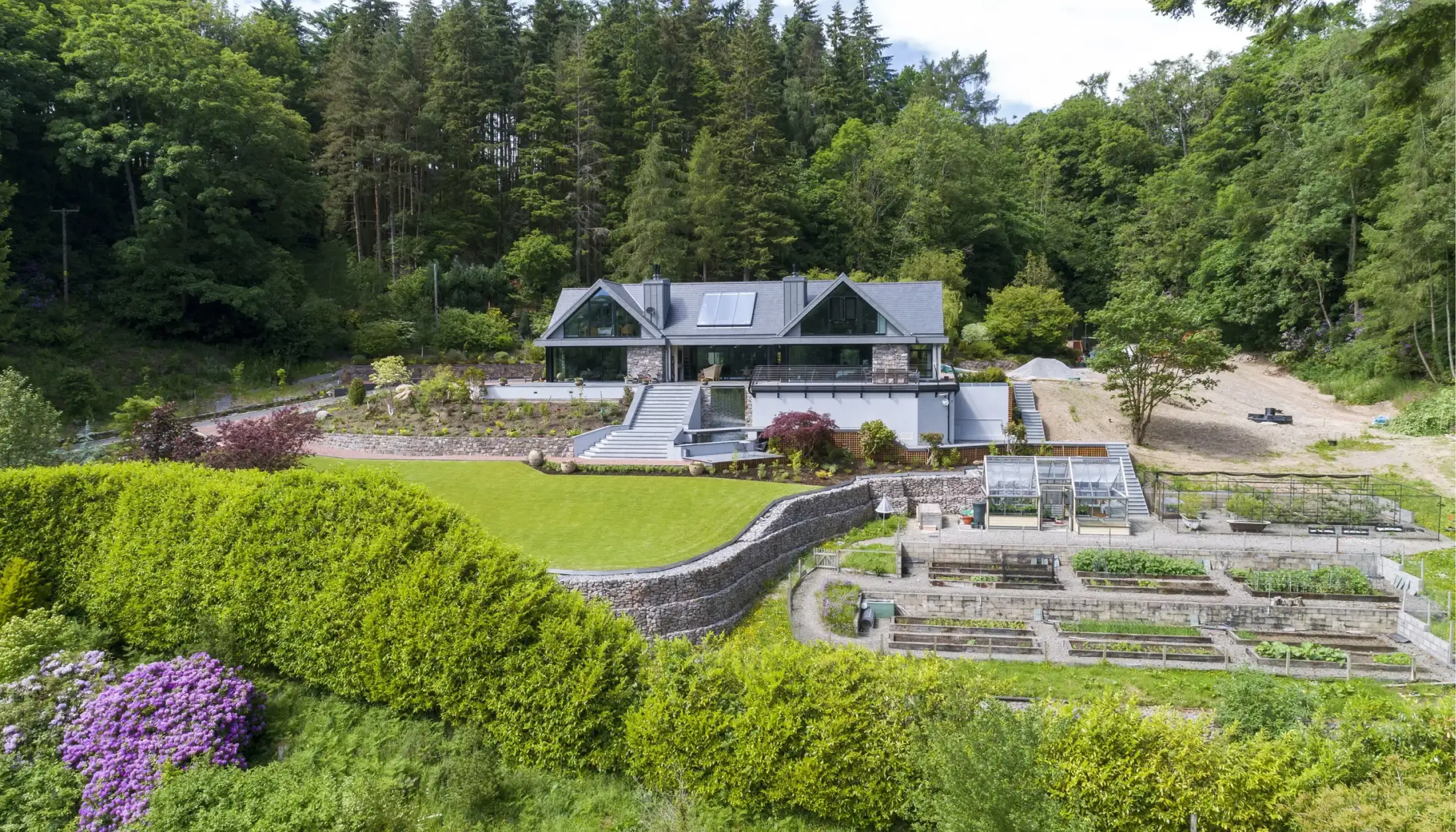 Aerial view of a grey-roofed house with black solar panels, surrounding green lawn on the right and gravel yard on the left.