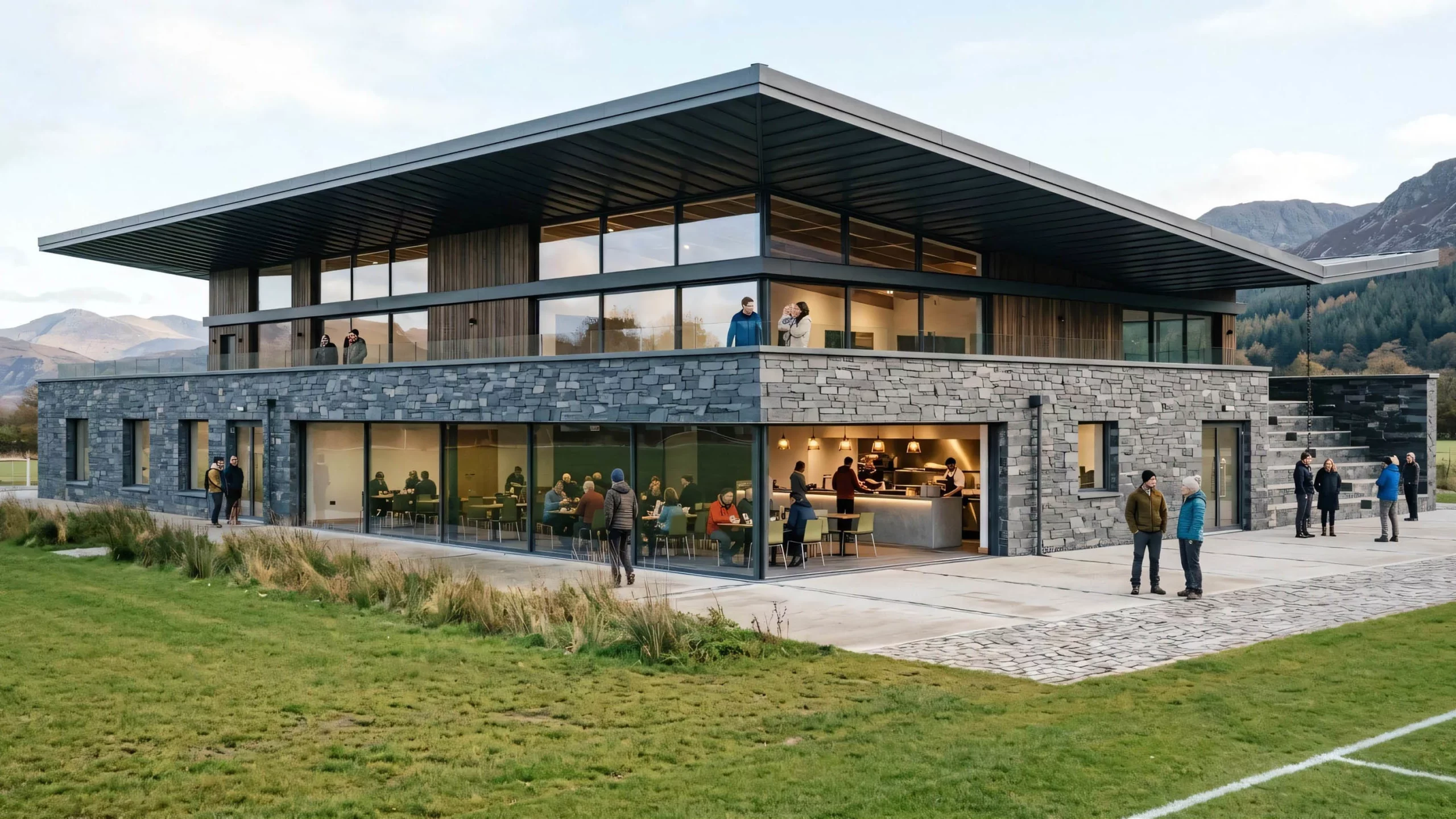 Modern two-storey clubhouse with stone lower walls and large glass windows showing people dining and standing on terrace, set by grassy sports pitch and hills.