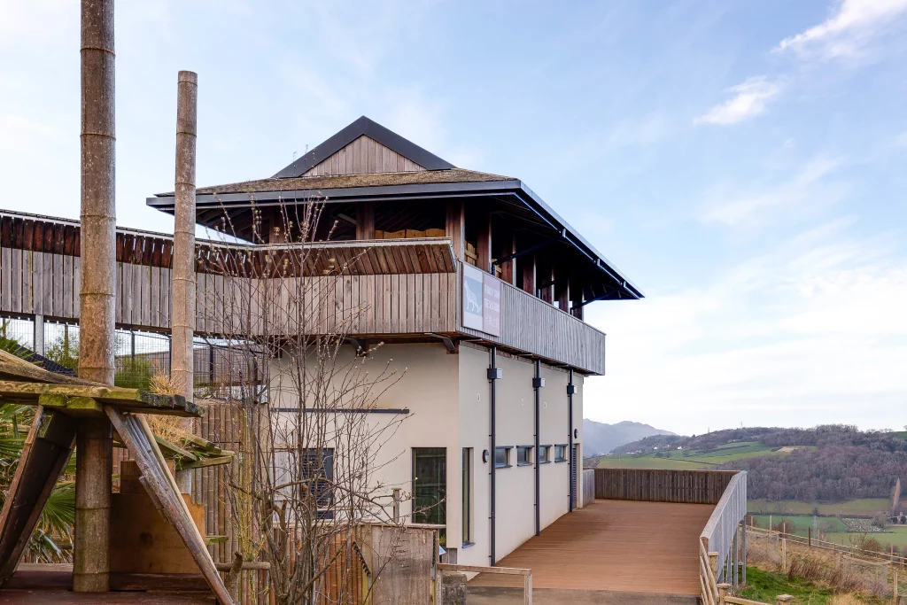 Wooden multi-level building with wide balcony and overhanging roof set against a clear sky and rolling hills background