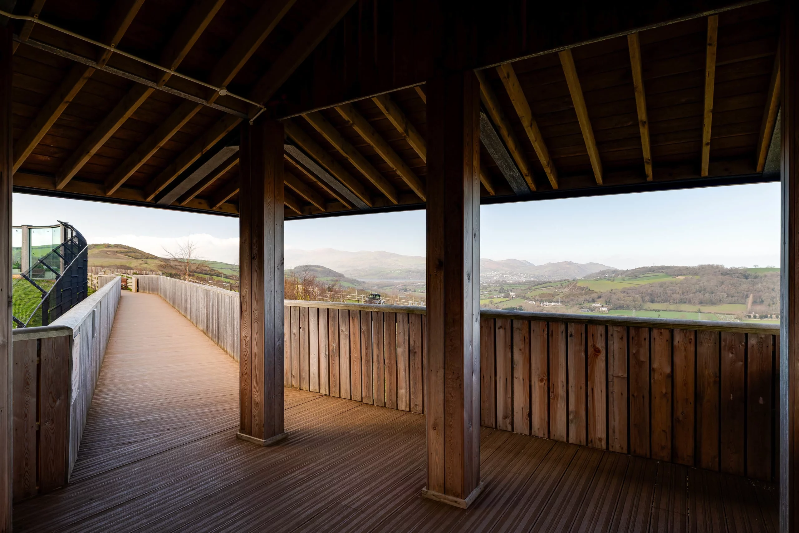 Covered wooden viewing platform with pillars framing a rolling green valley and distant hills through wide open windows