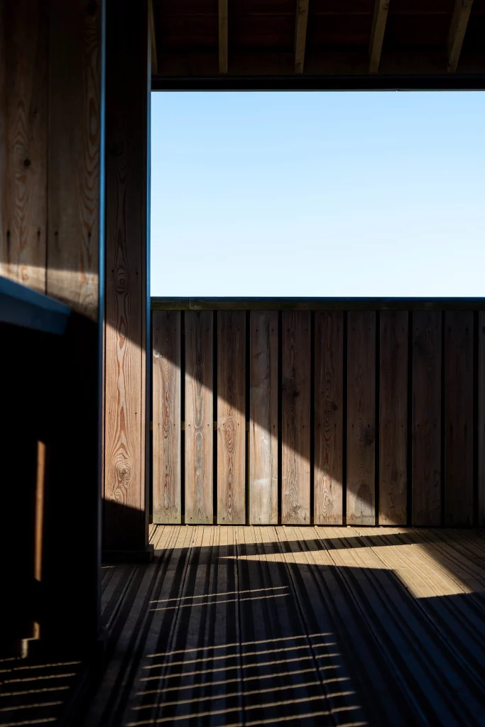 Sunlit wooden deck and vertical plank wall with strong diagonal shadows and a pale blue sky visible through a rectangular opening