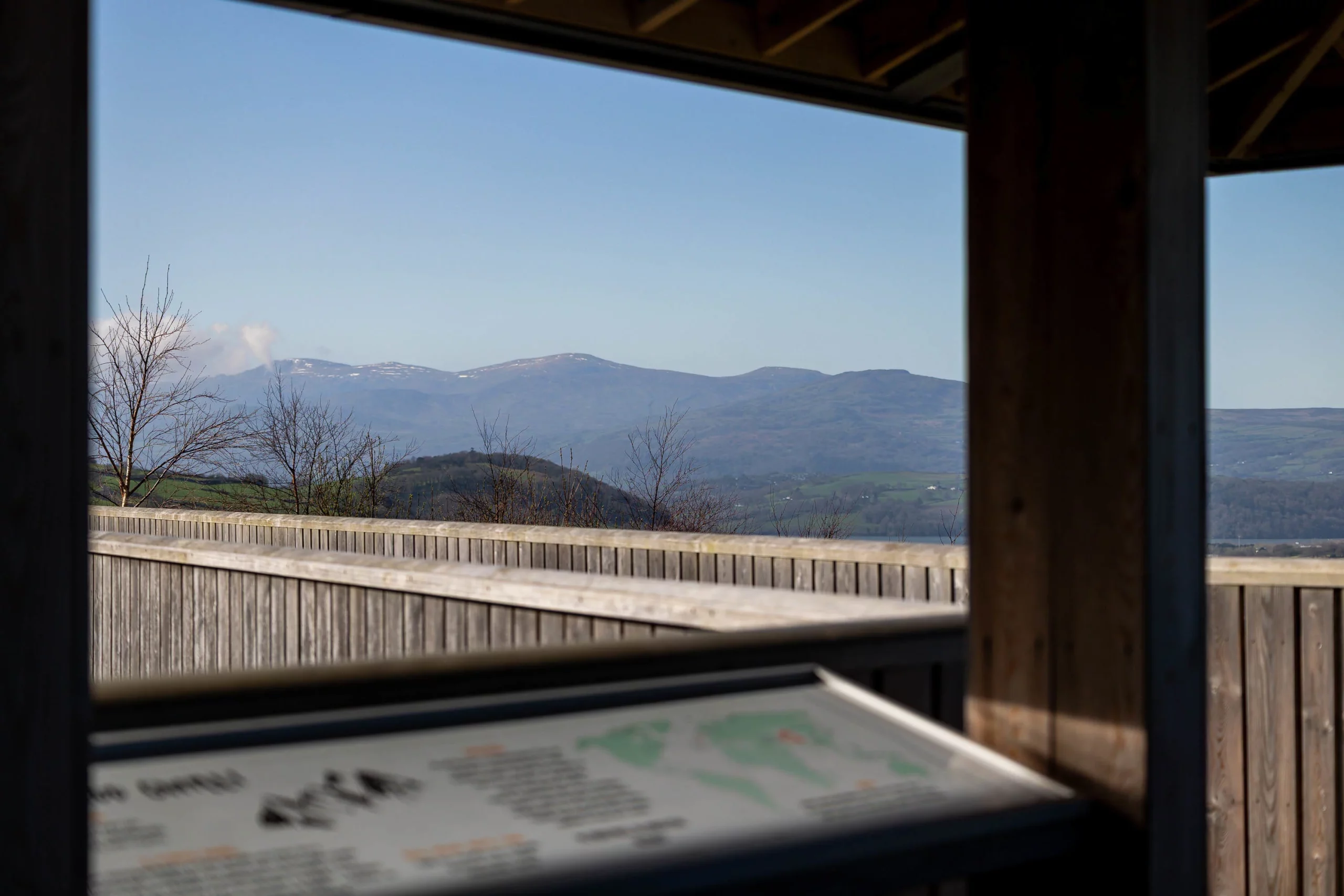 Wooden viewing platform framing distant snow-capped hills and bare trees under a clear blue sky.