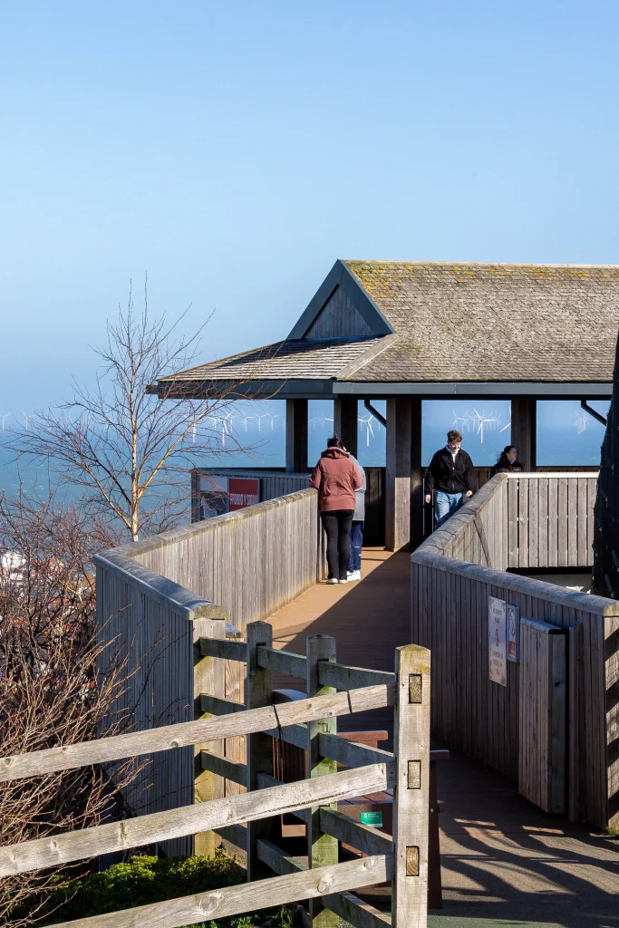Wooden seaside viewing platform with a sloped roof and people walking along the boardwalk, wind turbines visible offshore.