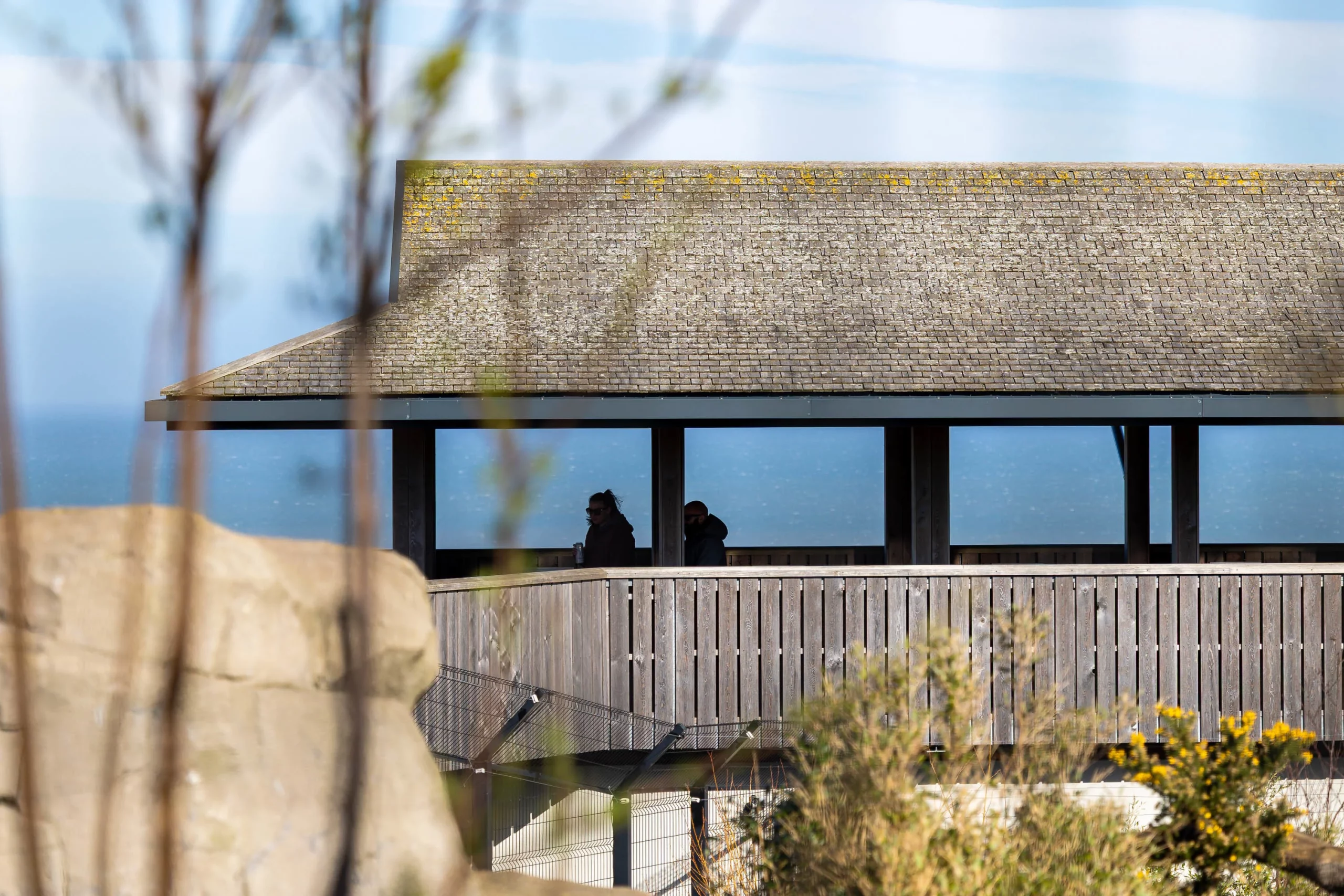 Wooden shelter with a weathered shingle roof and railing, two silhouetted people visible inside against a blue seascape background