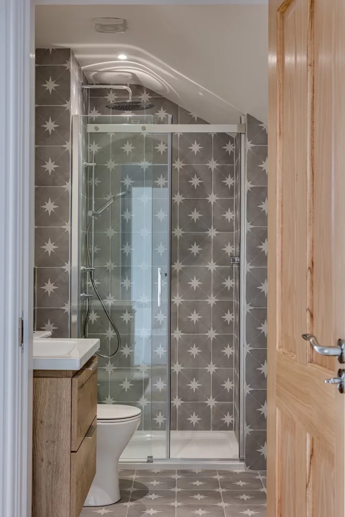 Small bathroom with glass shower enclosure, grey star-patterned tiles, white toilet and wooden vanity visible through open door.