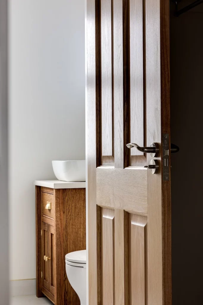 Partially open wooden bathroom door showing brass handle, glimpse of white toilet and wooden vanity with vessel sink.