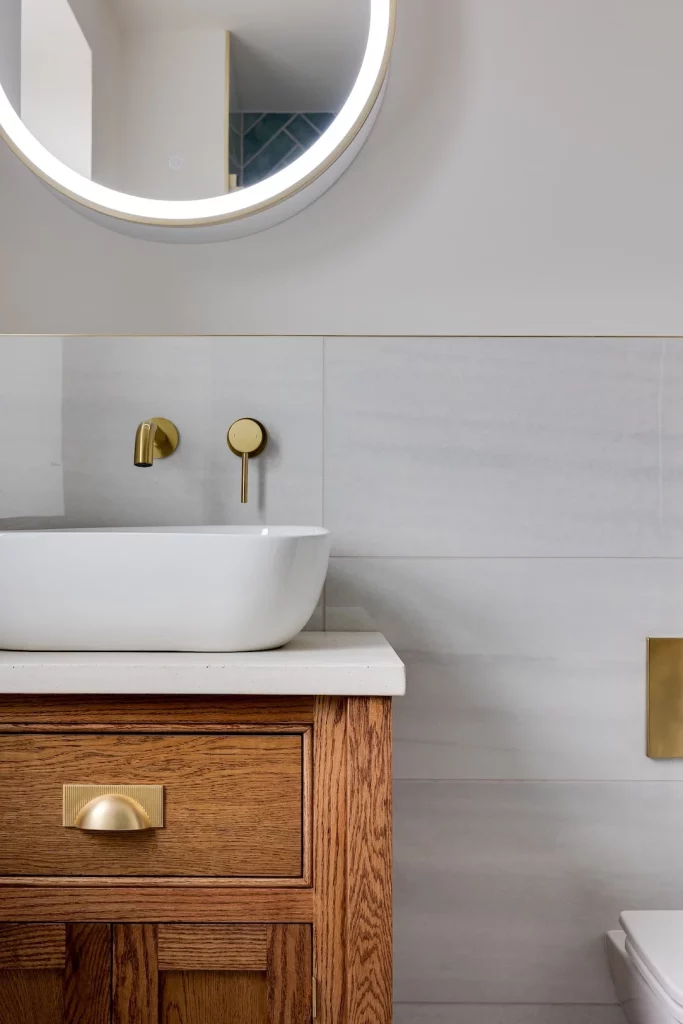 Round illuminated mirror above white vessel sink, brass wall tap and wooden vanity drawer with brass cup handle.