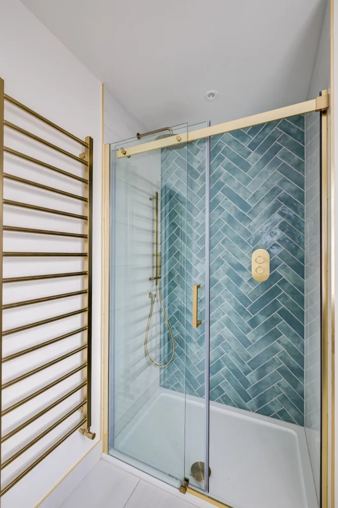 Shower with sliding glass door, gold hardware and handheld shower against blue-green herringbone tile, gold towel warmer nearby