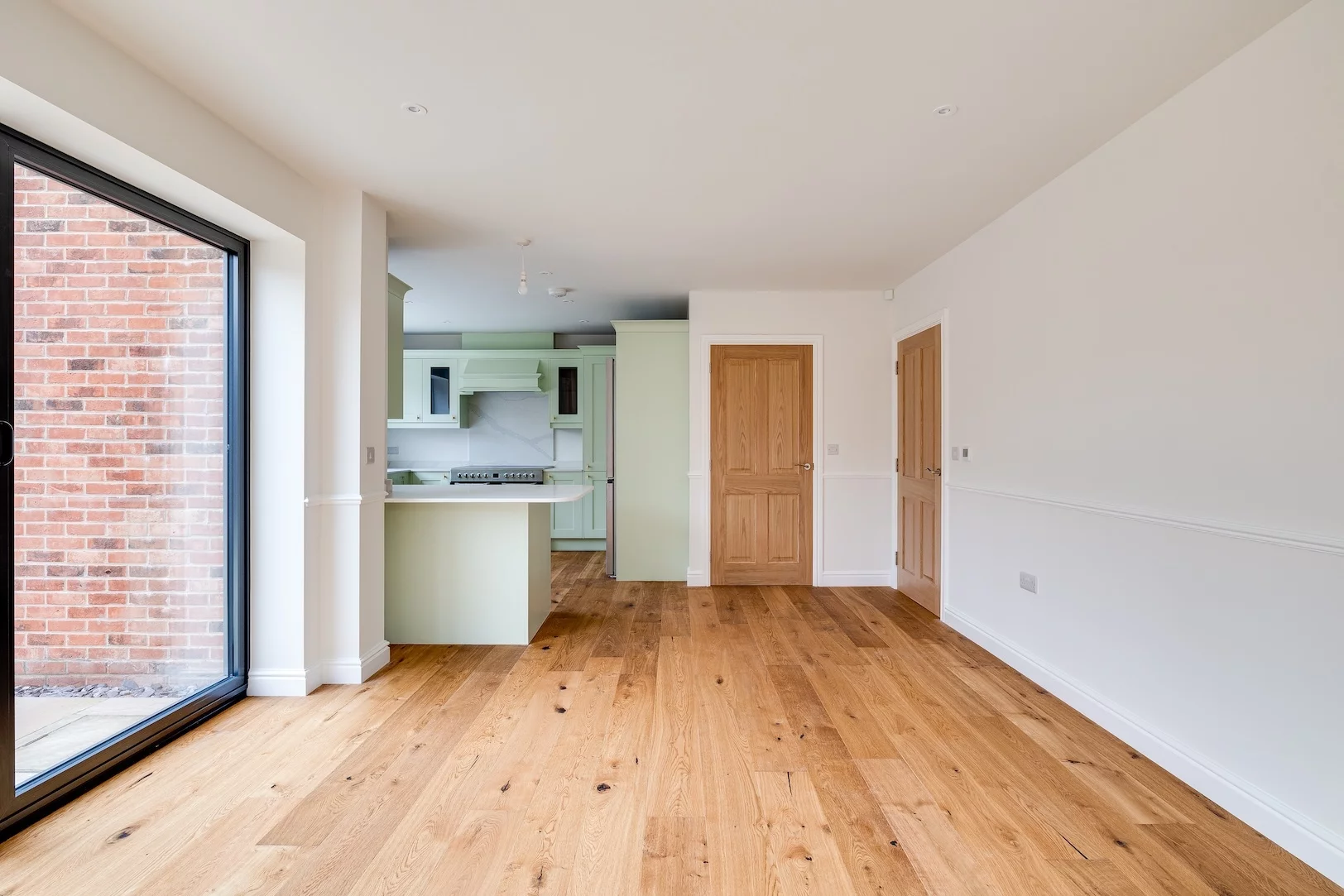 Spacious open-plan kitchen and dining area with pale green cabinets, wooden floor, two wooden doors and large sliding window to brick exterior.