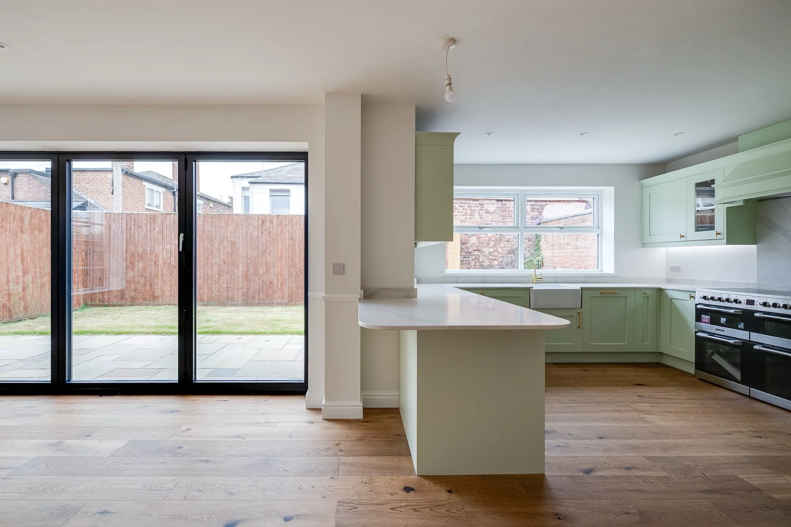 Open-plan kitchen with pale green cabinets, white countertops, farmhouse sink under a window, large black-framed sliding glass doors to a fenced backyard.