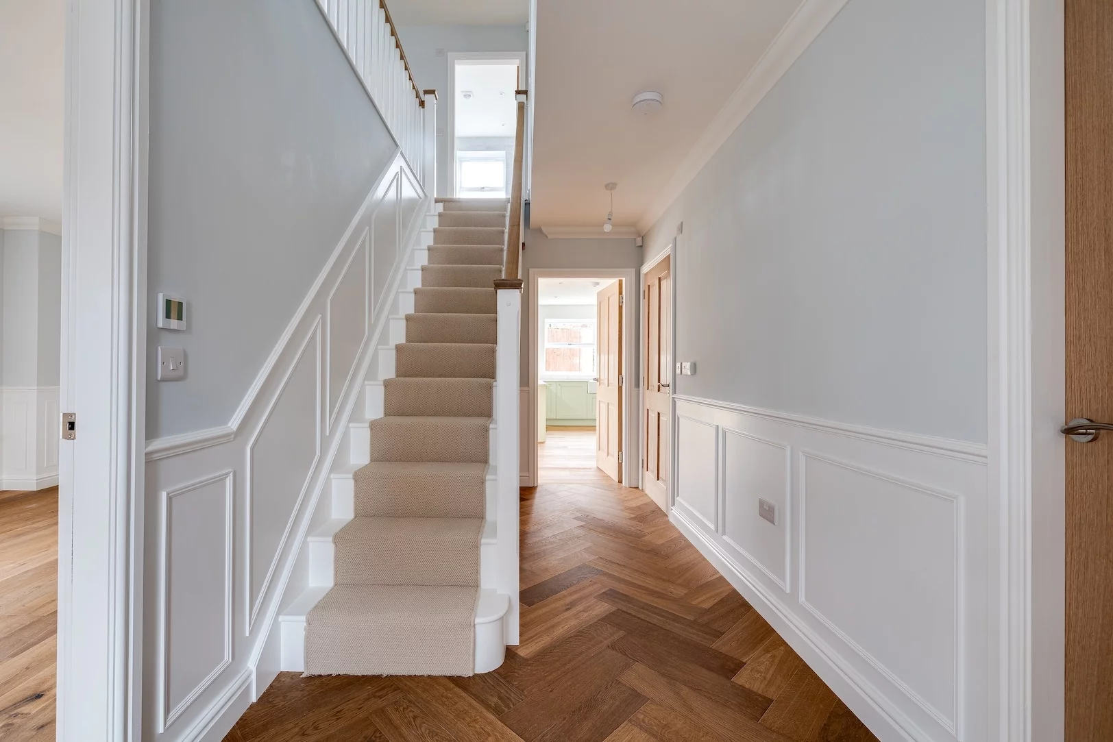 Bright hallway with carpeted central staircase, white panelling walls and herringbone wood floor leading to an open doorway.