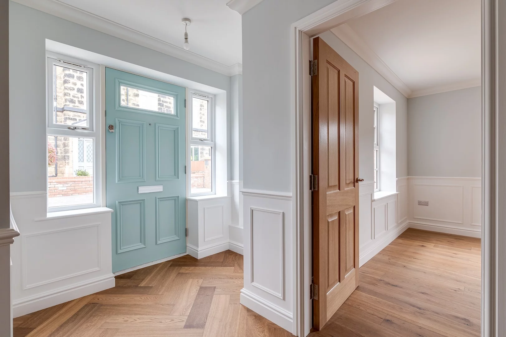 Hallway with pale blue paneled front door flanked by sidelights, white wainscoting and herringbone oak flooring, open wooden interior door.