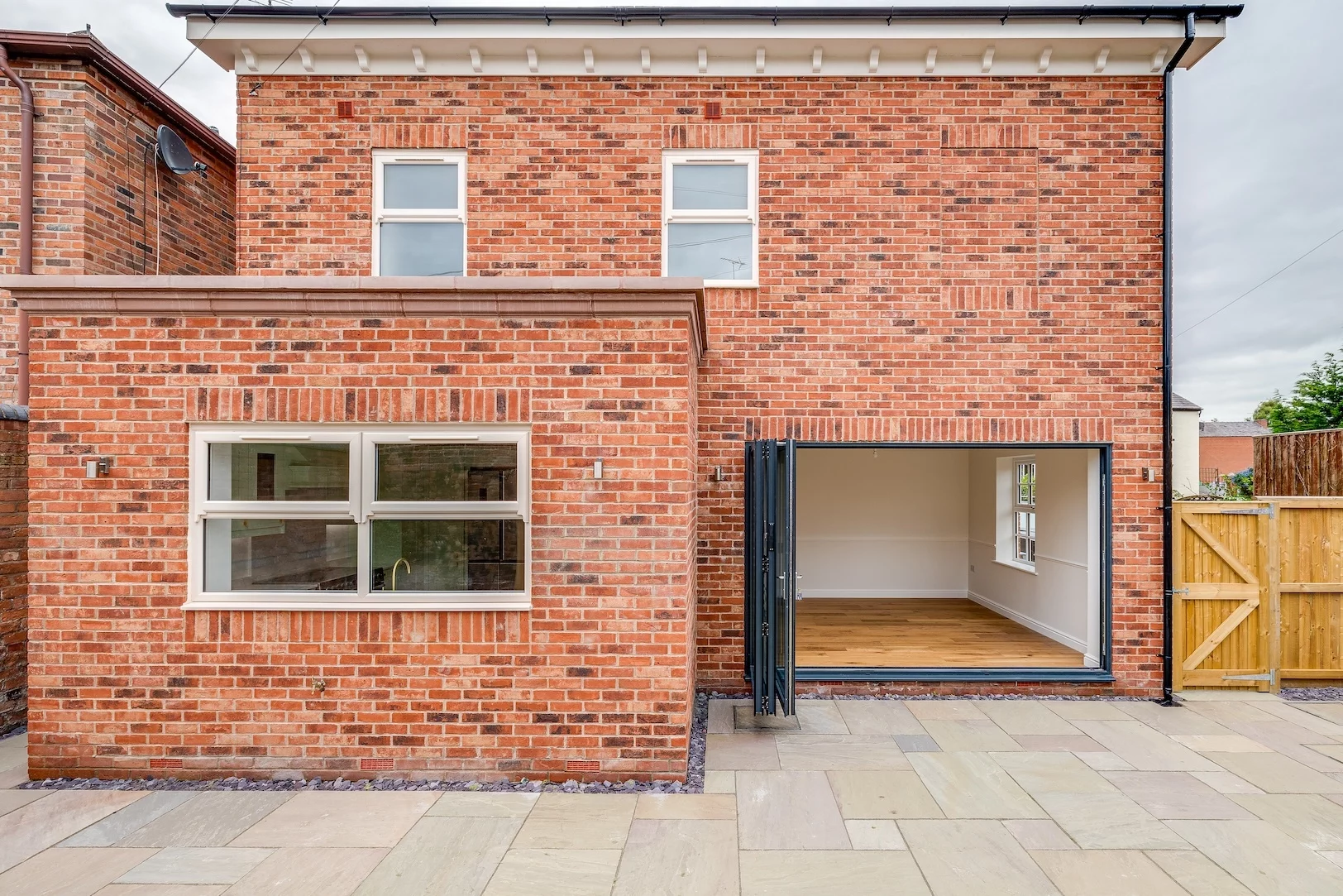 Rear view of a modern red-brick two-storey house with patio, a large open folding-glass door to a wooden-floored room and white-framed windows.