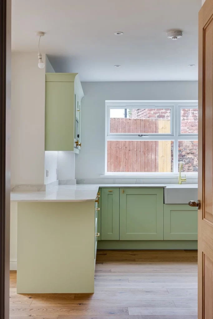 Compact kitchen with pale green shaker cabinets, white marble countertop, farmhouse sink and brass tap under a wide window showing a wooden fence.
