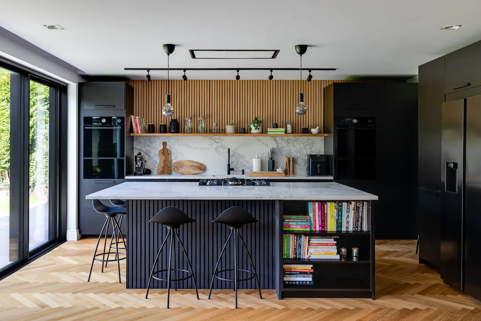 Modern kitchen with marble island, black bar stools and built-in bookshelf, fluted wood feature wall and pendant lights over a hob.