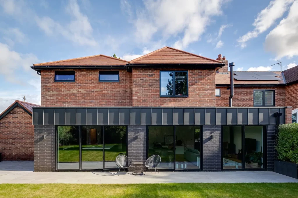 Two-storey red brick house with large black-framed sliding glass doors, patio with two wire chairs and a green lawn.