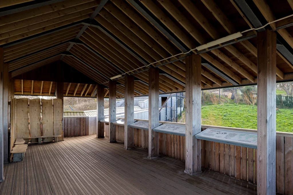 Open wooden shelter with angled roof beams, vertical posts and display panels overlooking grassy hillside.