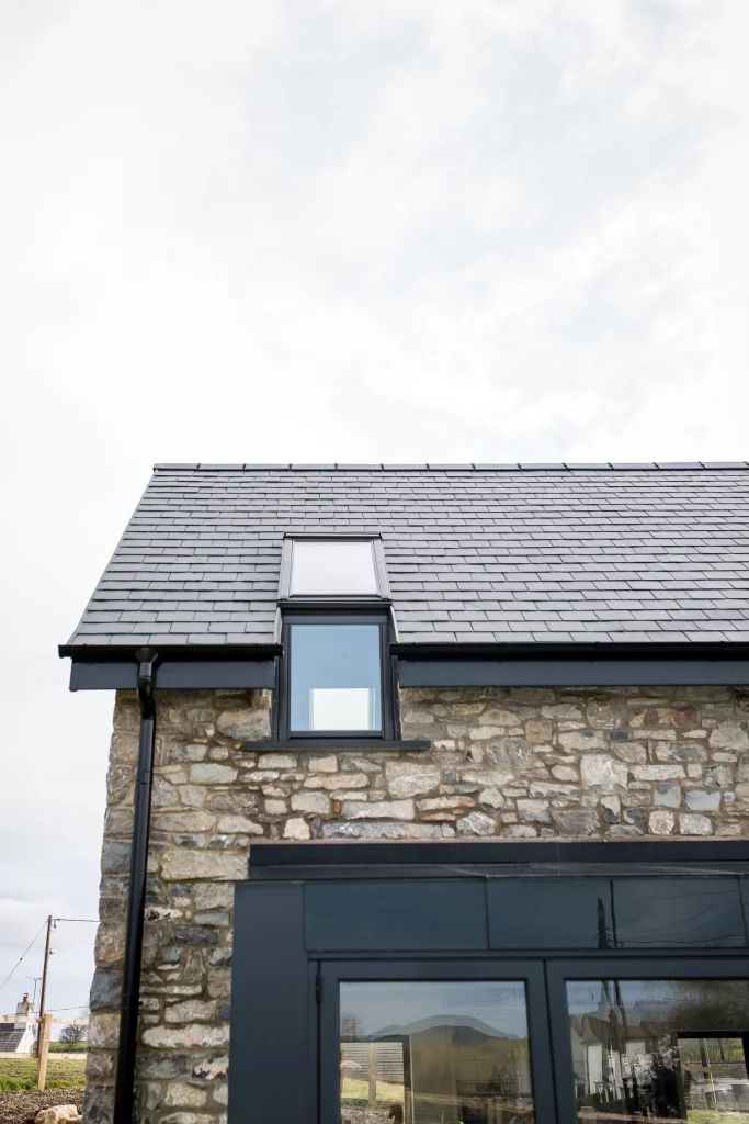 Slate-roofed stone house gable with black-framed dormer window and rain gutter under a pale cloudy sky