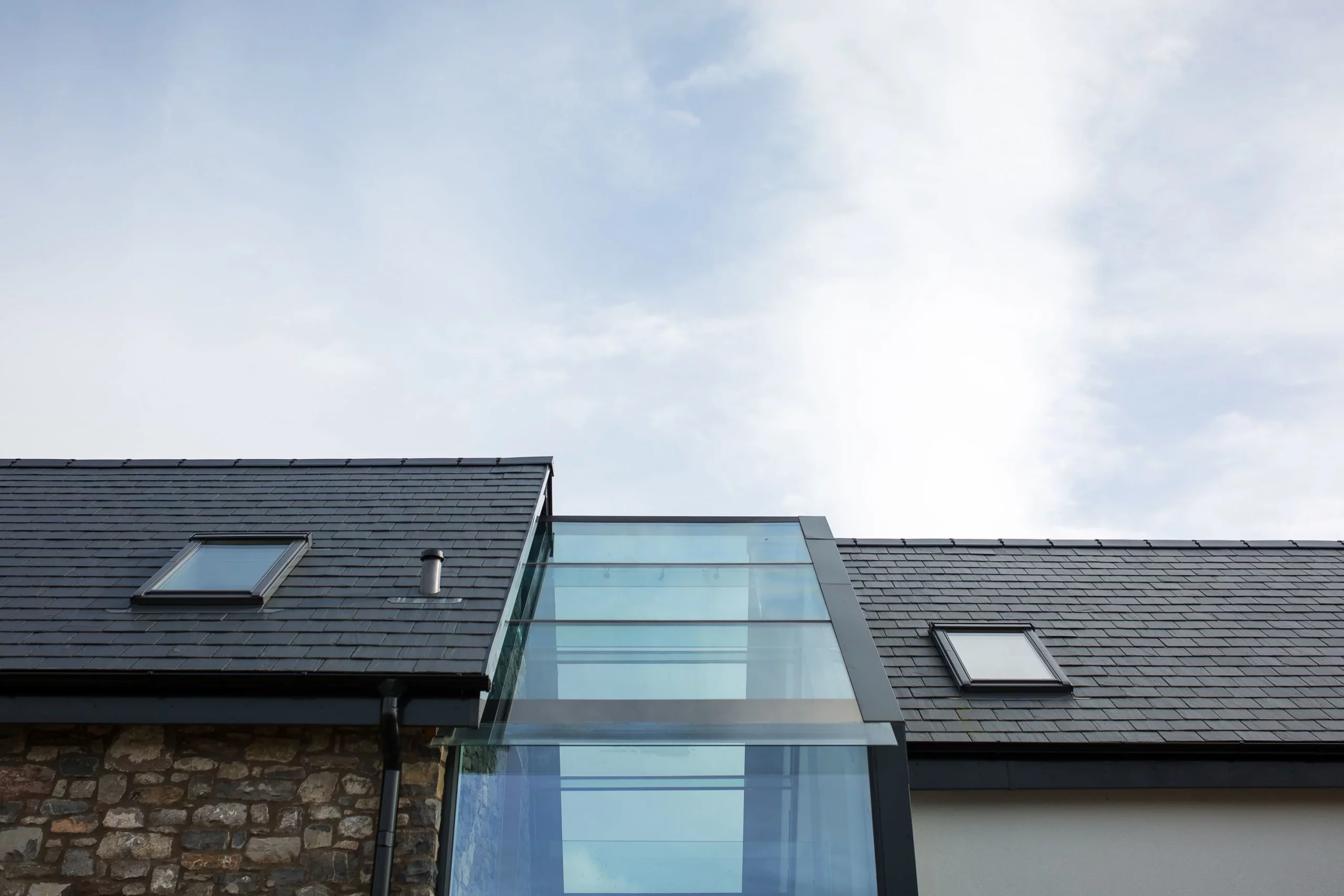 Slate-tiled roof with two skylights and a central glass atrium above a stone and rendered wall, under a pale cloudy sky