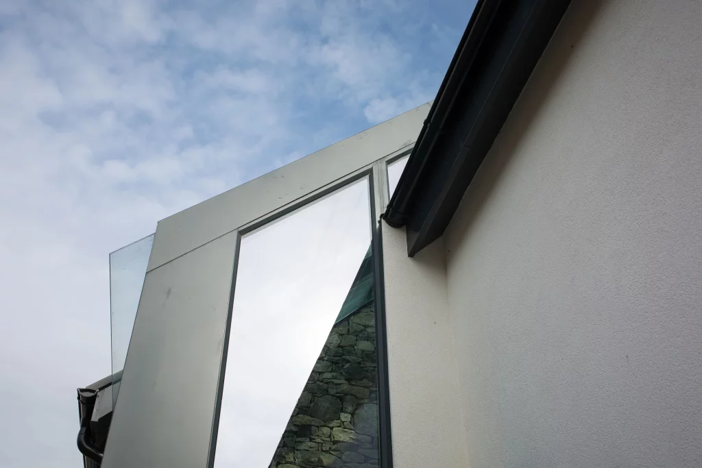 Angled close-up of a modern building corner showing glass panels, a reflective window, stonework reflection and a black gutter against a pale wall