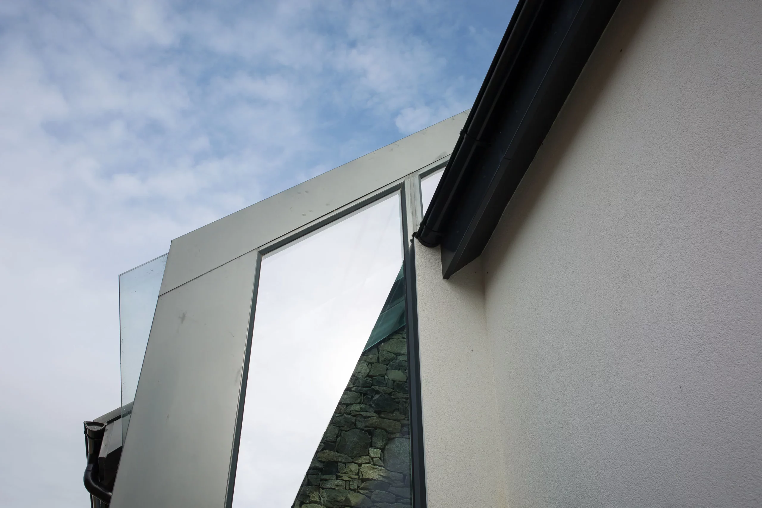Angled close-up of a modern building corner showing glass panels, a reflective window, stonework reflection and a black gutter against a pale wall