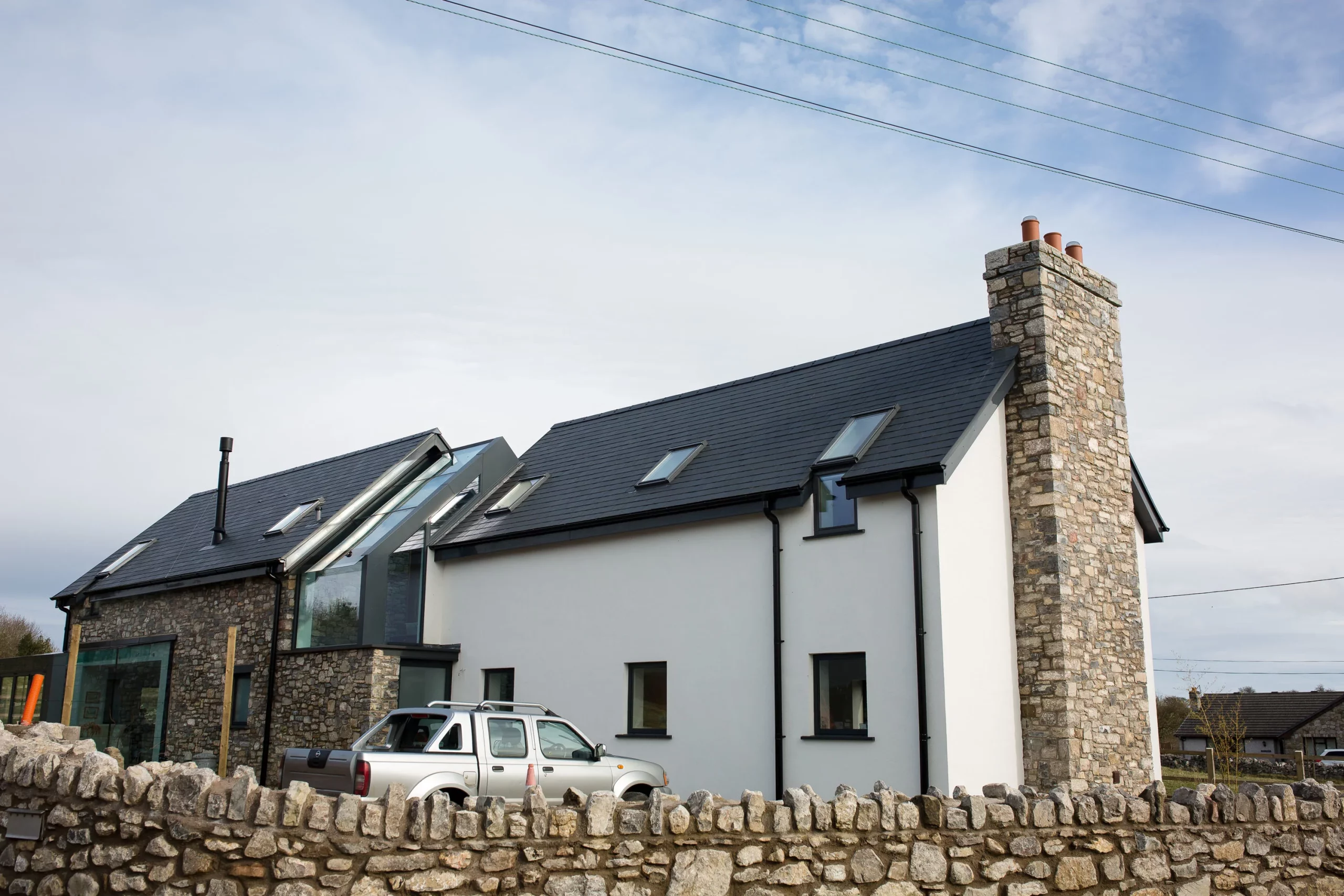 Modern stone-and-plaster house with slate roof, skylights and tall stone chimney behind a low dry-stone wall, silver pickup parked outside.