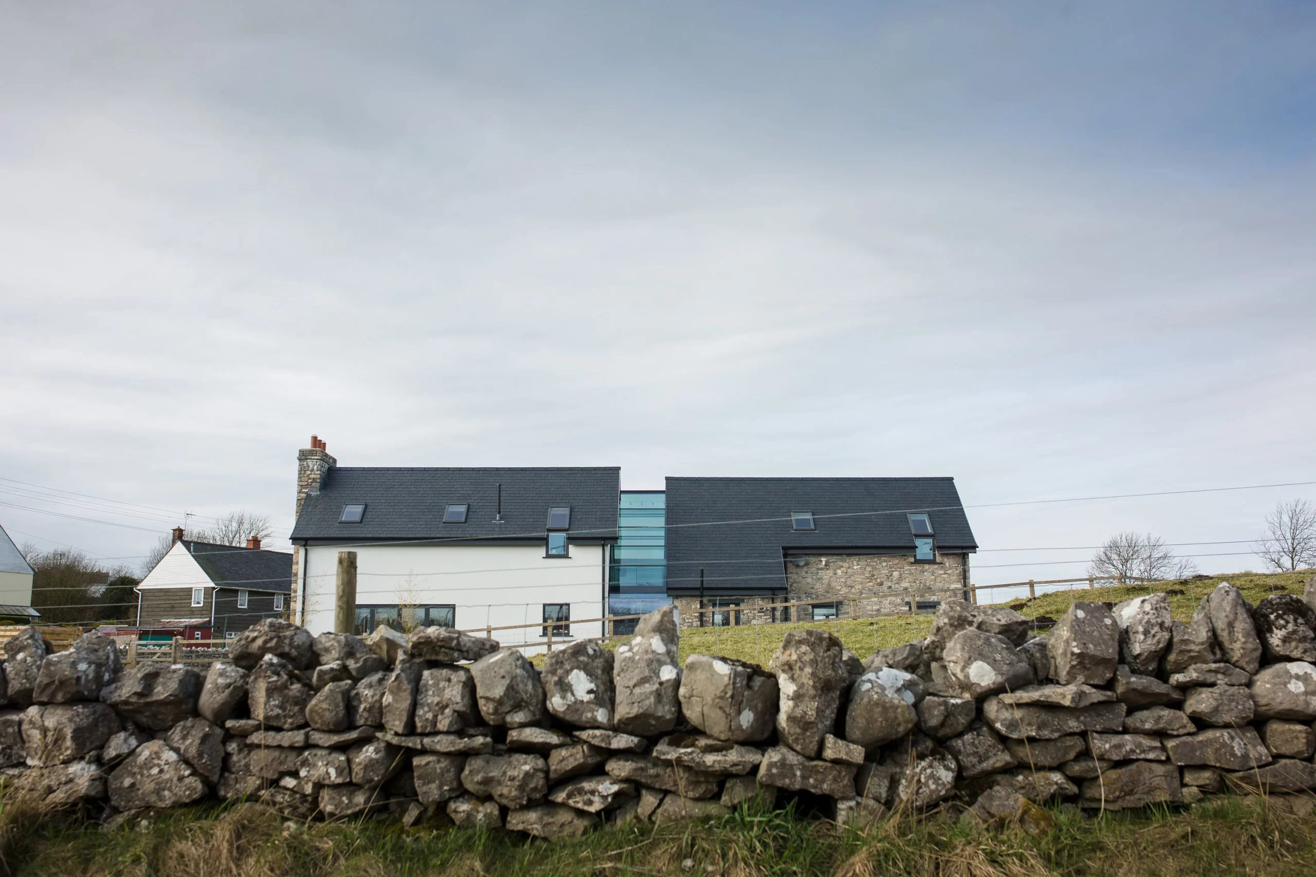 Modern slate-roofed farmhouse with stone lower walls behind a dry-stone wall in a grassy rural setting