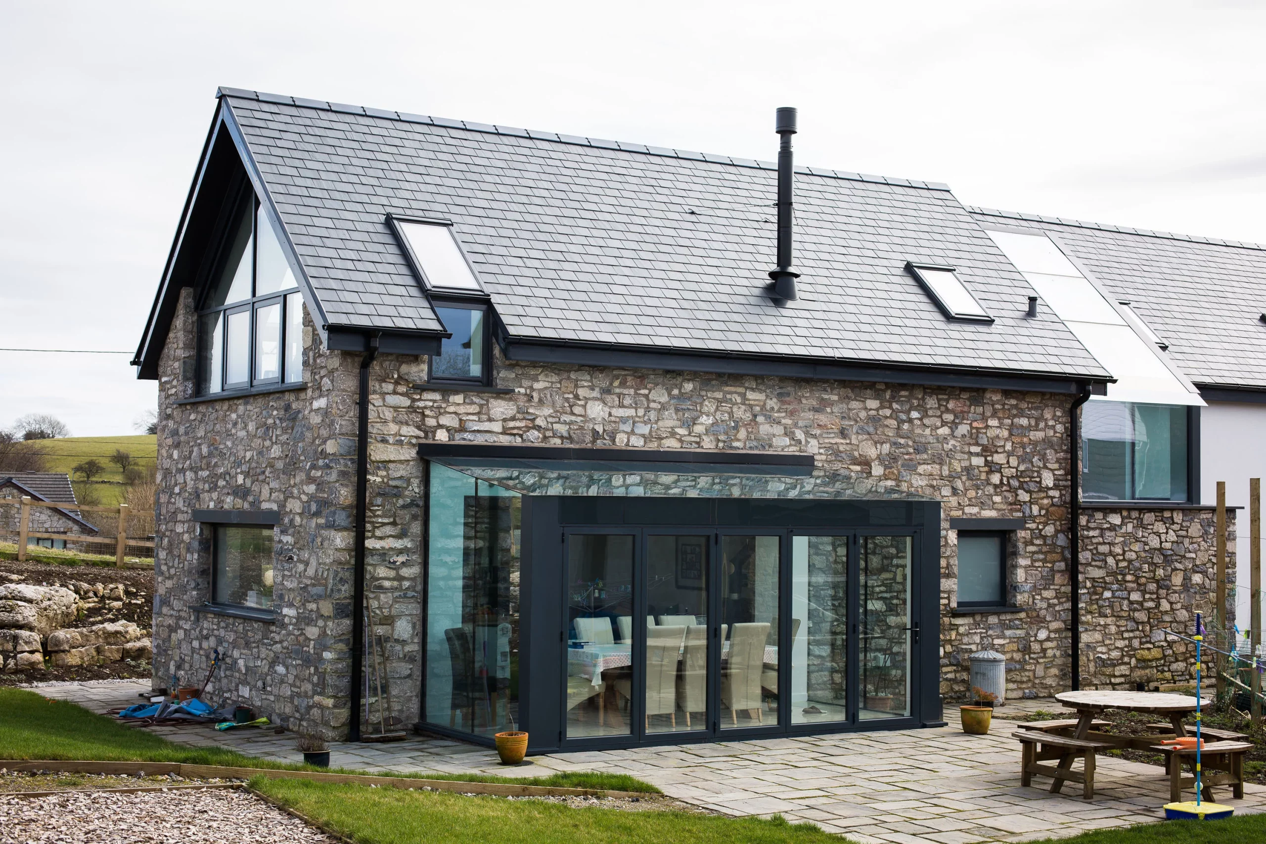Stone cottage with slate roof, roof windows and tall black chimney, glass-walled dining extension and patio furniture