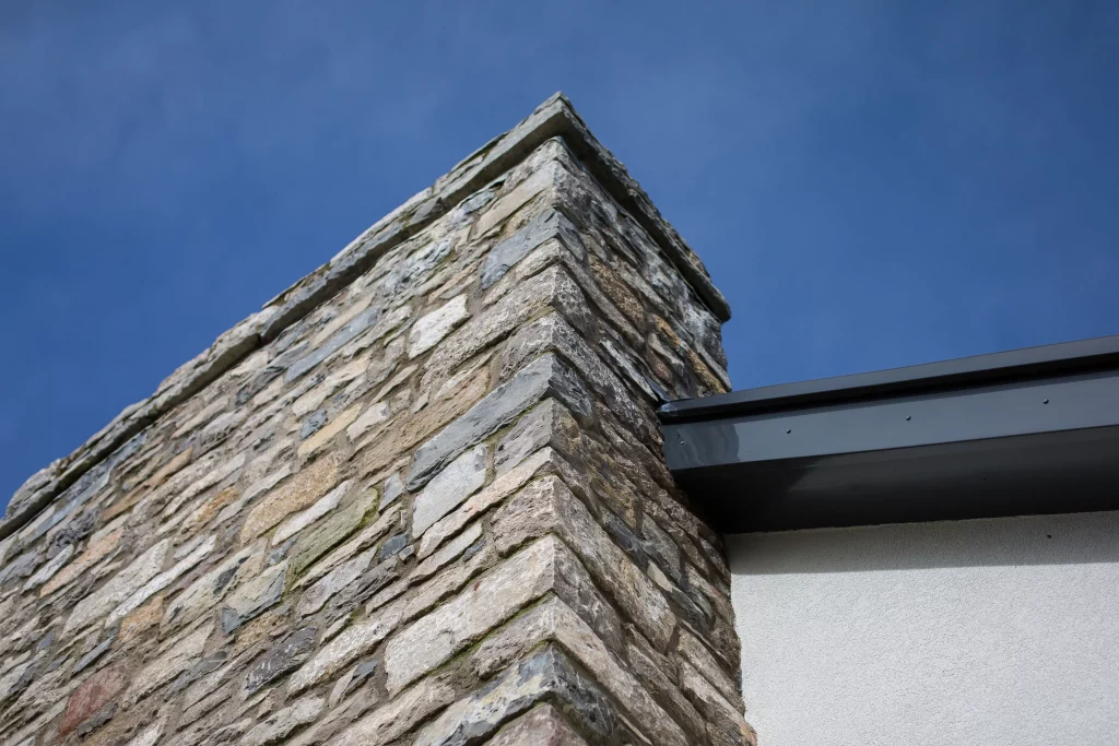 Corner of a stone-clad chimney rising against a clear blue sky with a metal roof edge and white rendered wall visible.