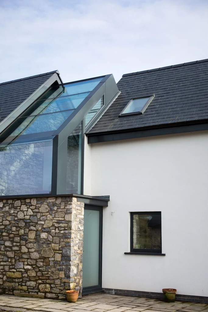 Modern house corner showing stone wall, white render, glass atrium roof and dark tiled sloped roof with a small skylight