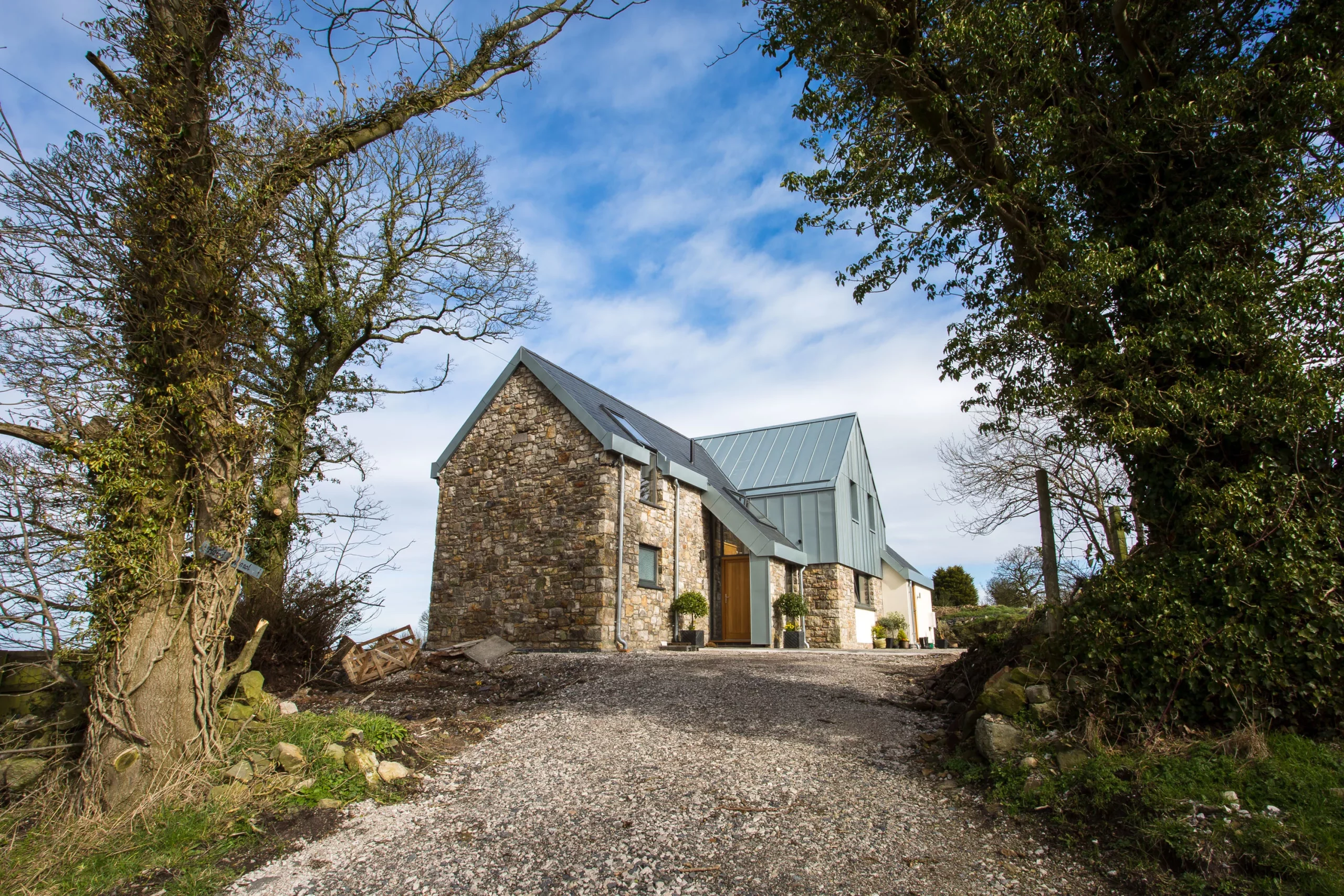 Stone cottage with modern metal roof and wooden door at the end of a gravel drive, framed by large trees and blue sky