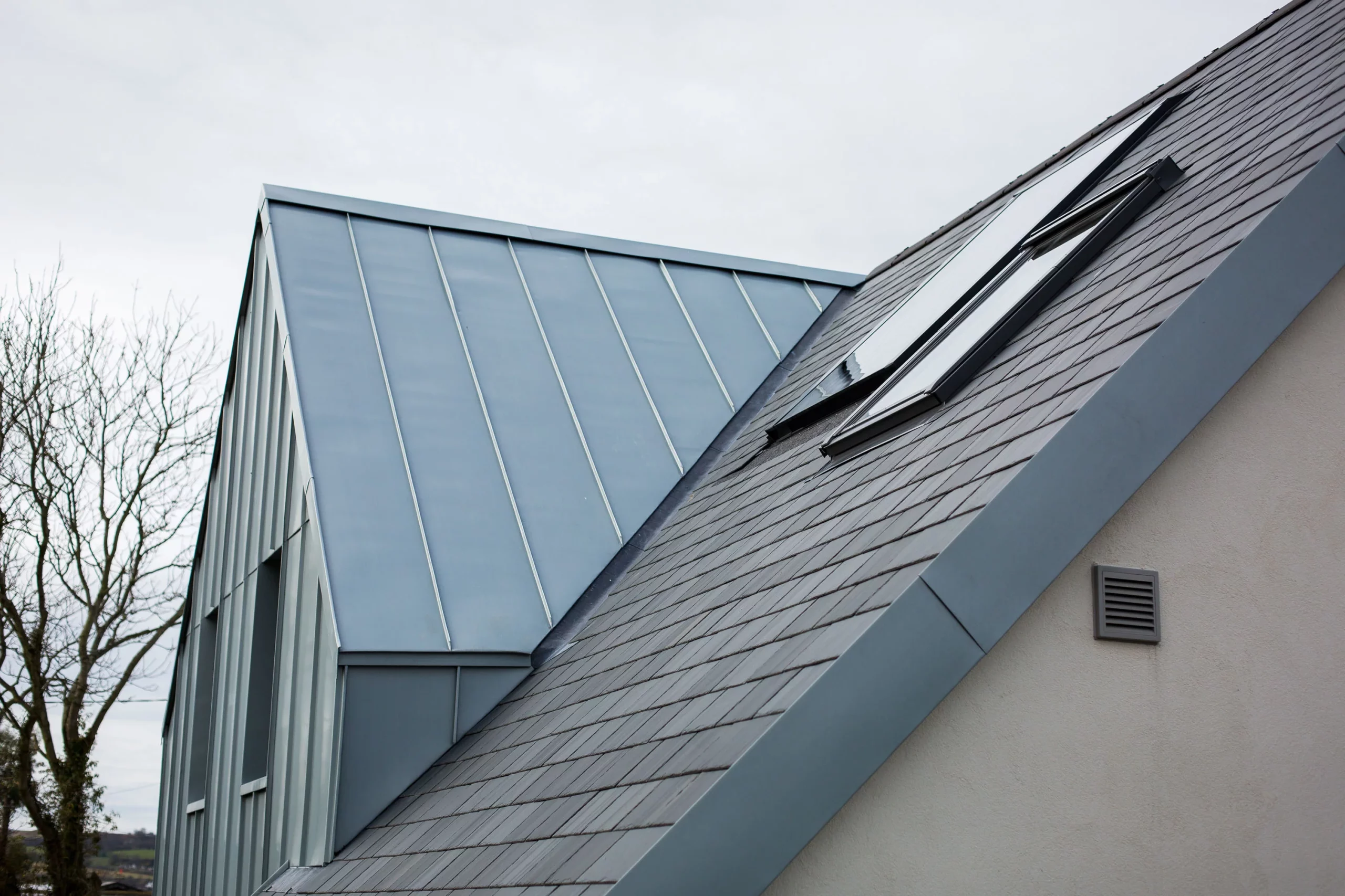 Slate-tiled pitched roof with two skylights meeting a standing-seam metal roof on a modern house, bare trees in background