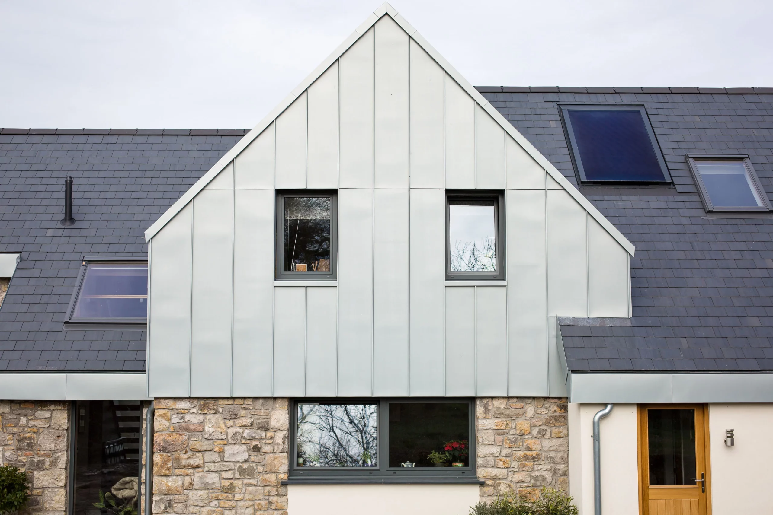 Modern house front with stone lower wall, pale metal gabled cladding, two small upper windows and dark slate roof with skylights.