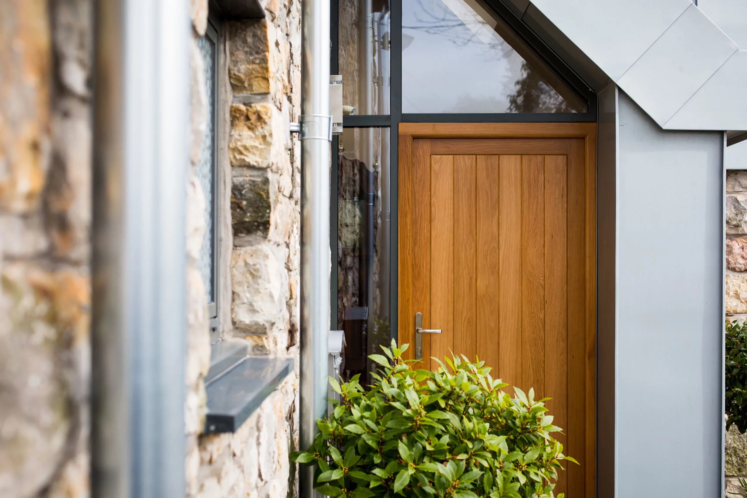 Vertical oak front door with metal handle set in a modern porch next to stone wall and green shrubbery