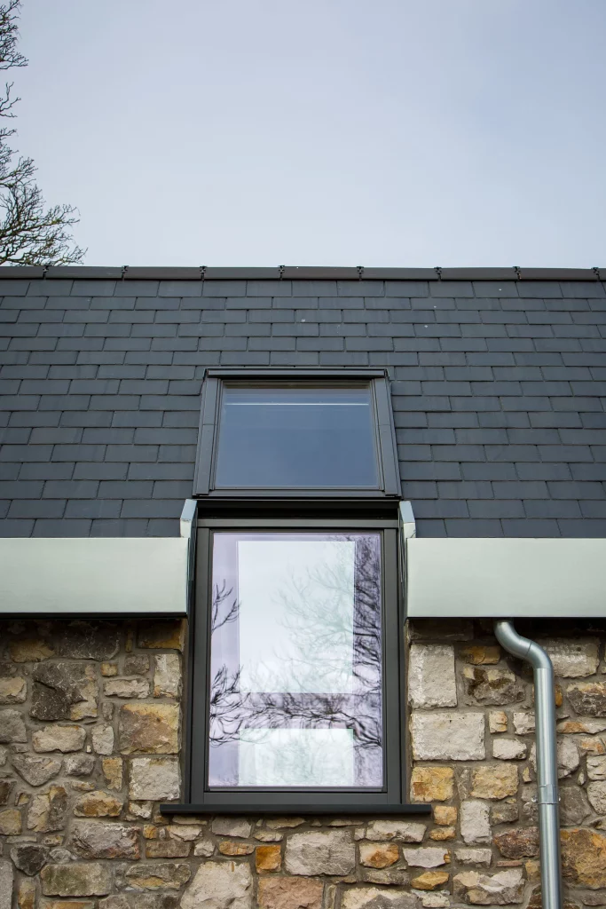 Tall narrow window set in stone wall with slate roof above and metal guttering, reflections of tree branches in the glass